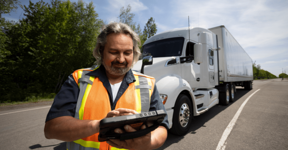 Truck Driver Using Tablet Roadside Truck driver wearing a safety vest using a tablet roadside with a white semi-truck in the background.